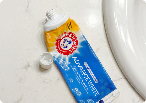Used ARM & HAMMER Advance White toothpaste tube with cap removed, lying on a bathroom countertop beside a sink.